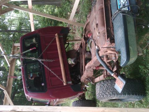 1992 Ford F-150 - A red pickup truck cab and its rusty frame are suspended by wooden beams and chains, undergoing repairs or disassembly.
