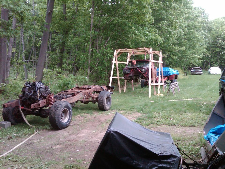 Rusty truck chassis with engine, another truck body on a wooden frame, and two cars in the background