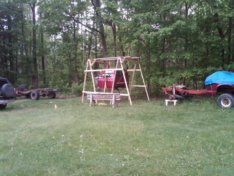 1992 Ford F-150 - A red truck cab is suspended in a wooden frame in a yard with other vehicle parts visible.
