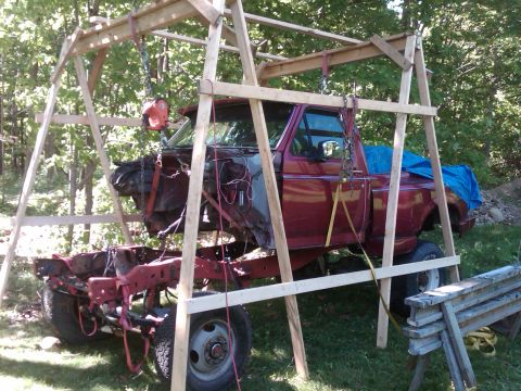 1992 Ford F-150 - Maroon Ford pickup truck body lifted by straps in a wooden A-frame gantry, separated from its rolling chassis in a wooded yard.