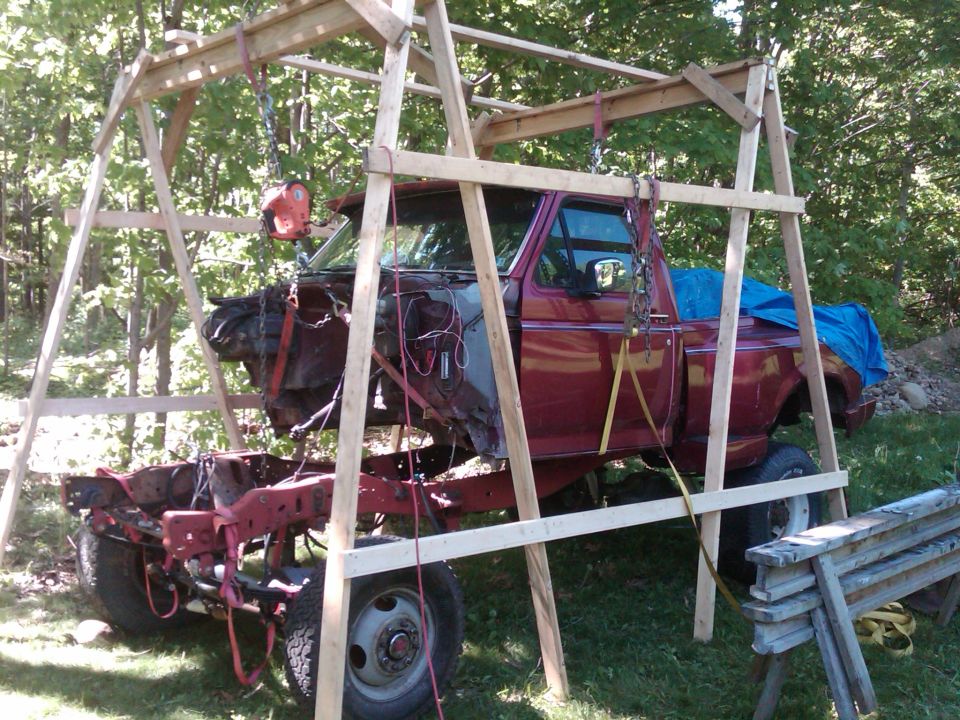 Maroon Ford pickup truck cab suspended in wooden A-frame gantry over its frame