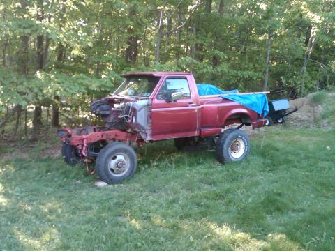 1992 Ford F-150 - A red pickup truck chassis sits in a grassy area, with the front end disassembled and the bed covered by a blue tarp.
