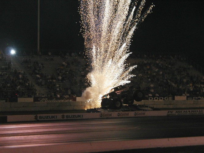Monster truck crushing cars at night on a drag strip with pyrotechnic sparks behind it