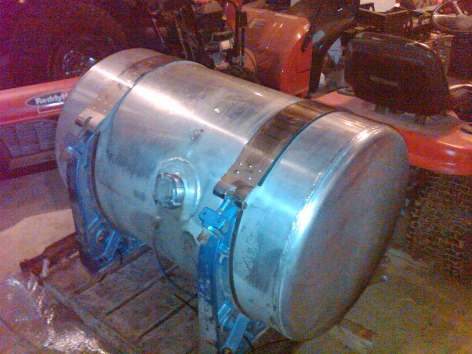large metal fuel tank with blue mounting brackets, sitting on a pallet. Reddy logo visible on red equipment in background