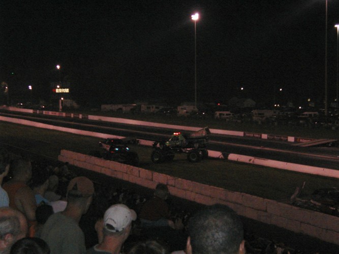 Monster truck and drag car on a drag strip at night, with spectators in the foreground
