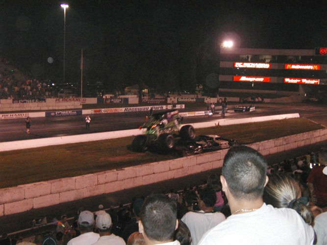 Monster truck crushing junk cars at night on a drag strip seen over a crowd