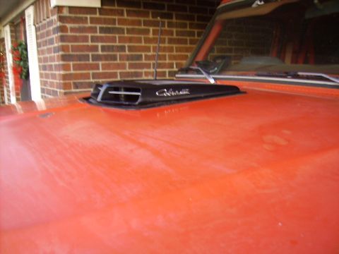 1978 Ford Bronco - Close-up of a black cowl-induction hood scoop with chrome script mounted on the orange hood of a pickup under a carport.