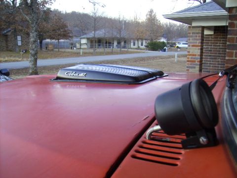 1978 Ford Bronco - Looking forward across a red-orange hood fitted with a black Cobra Jet-lettered hood scoop and a set of louvered vents; a round black auxil…