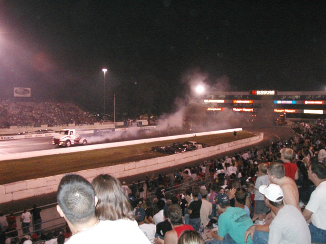 White and red truck doing a burnout on a drag strip at night, with spectators in the foreground