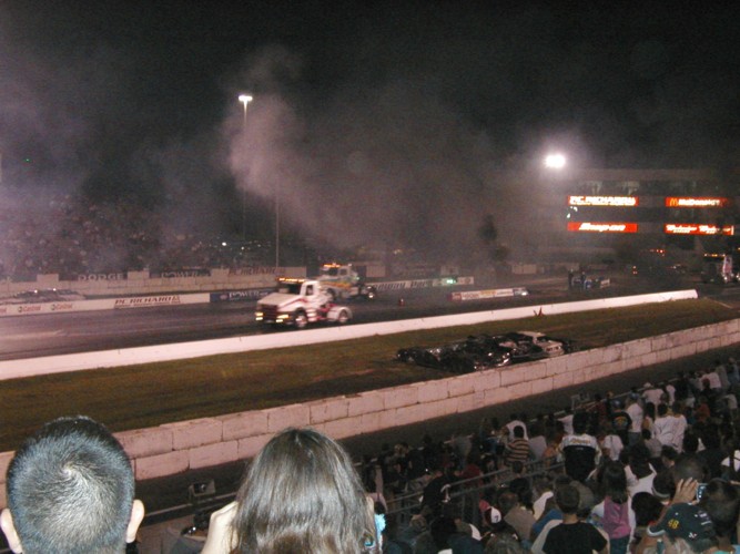 Two trucks race at night, one emitting smoke