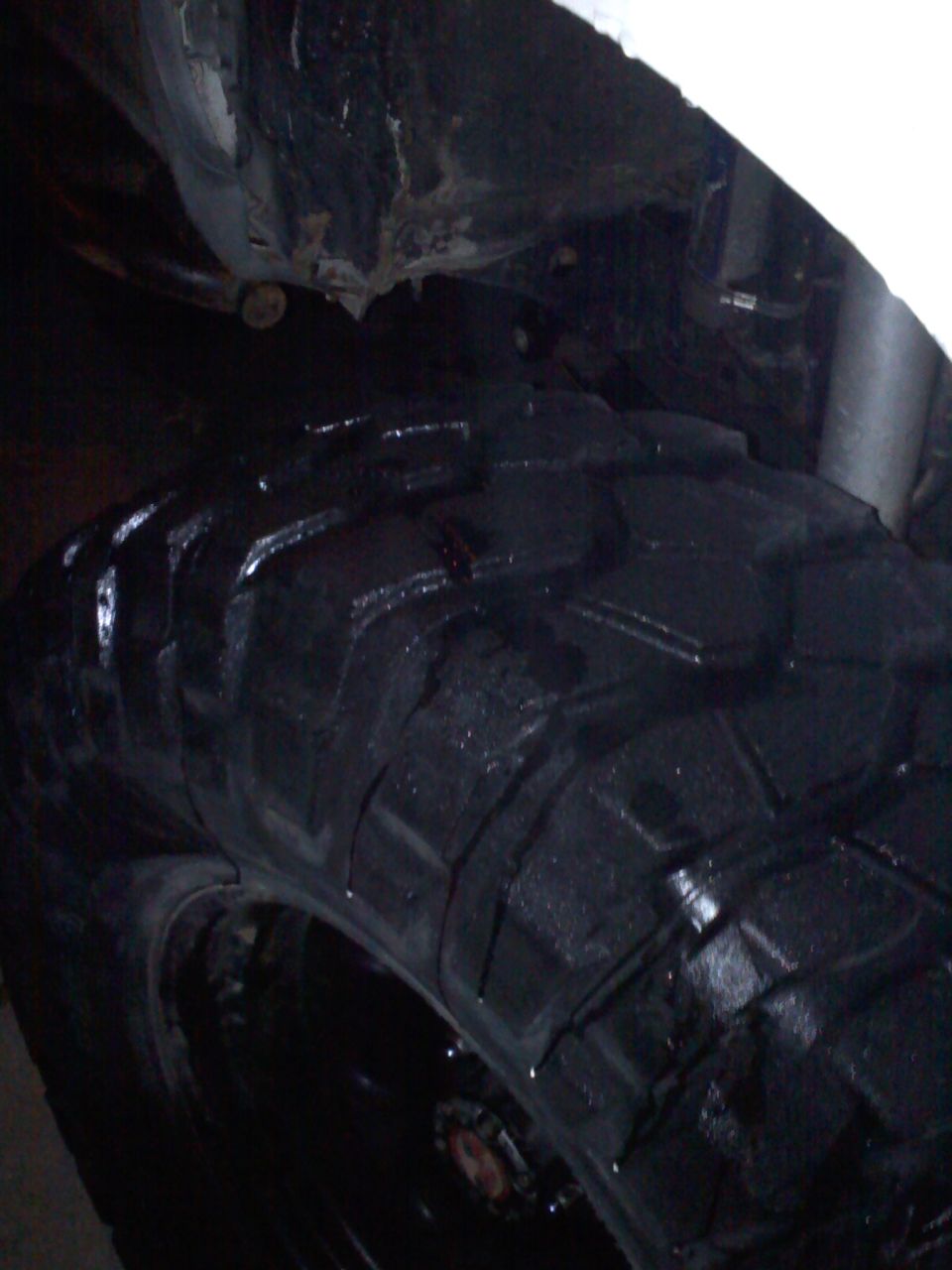 Black off-road tire with aggressive tread pattern, wet surface, and visible undercarriage components