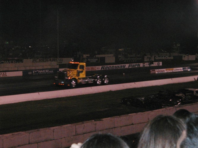 Yellow semi-truck with chrome wheels on a race track at night, another car visible in the distance