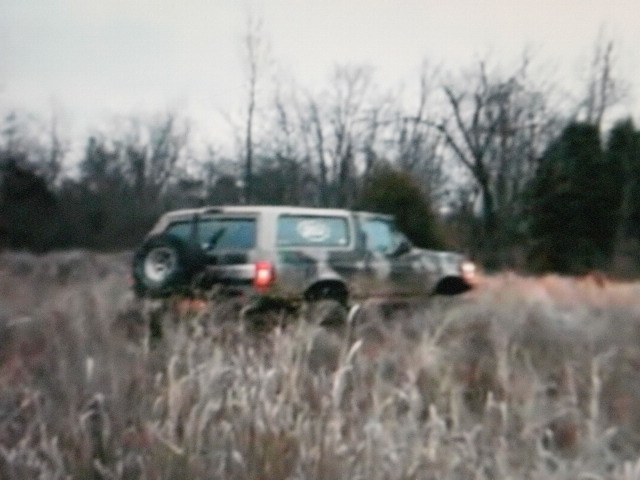 1995 Ford Bronco driving through tall dry grass with trees in background