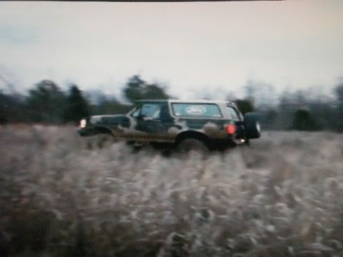 1995 Ford Bronco - A Ford Bronco is shown driving through a grassy field with trees in the background.