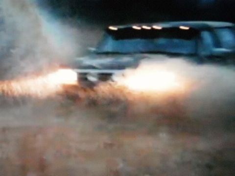1995 Ford Bronco - A dark blue Ford Bronco navigates through murky water, its headlights cutting through the spray.
