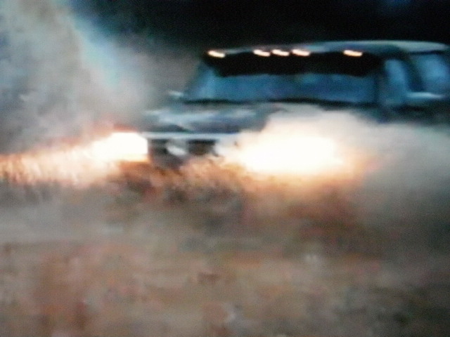 Dark blue 1992 Ford Bronco driving through water, headlights on, with roof lights illuminated