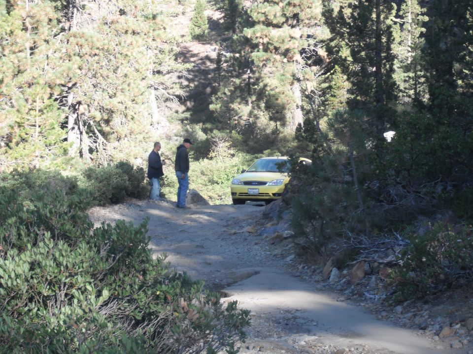 Yellow sedan parked on a rough dirt road in a wooded area with two people standing nearby