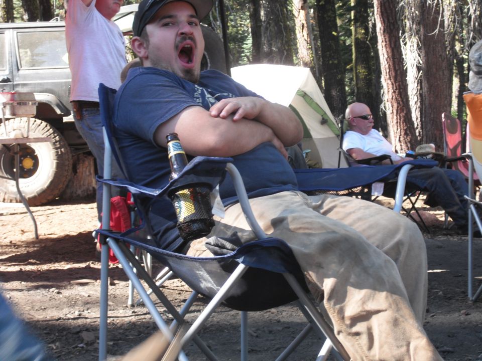 Man yawning in a camping chair with a beer, other people and tents in the background