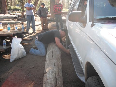 1991 Ford Bronco - A man kneels to work on the wheel of a white SUV, which is positioned next to a large log.