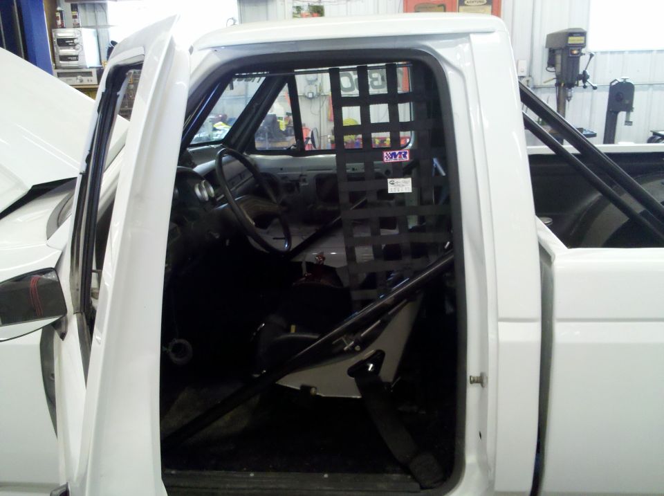 Open door of a stripped white race pickup showing roll cage door bars and black window net