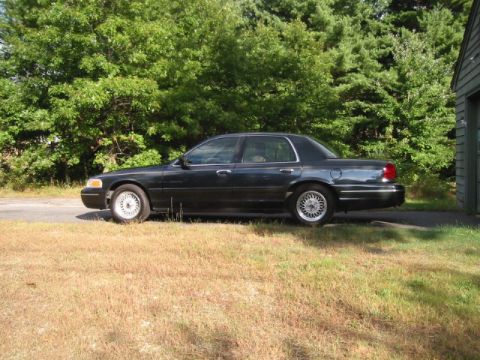1998 Ford Crown Victoria - A dark-colored 1998 Ford Crown Victoria is parked on a paved surface with dry grass in the foreground and trees in the background.