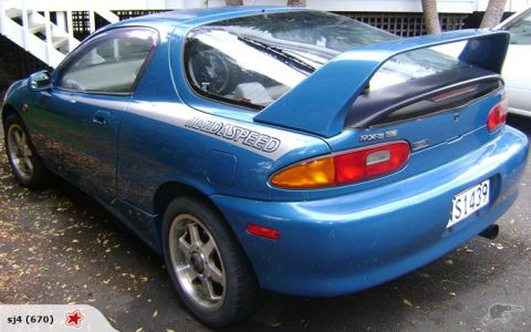 1993 Mazda MX-3 - A blue 1993 Mazda MX-3 is parked outdoors, showing its rear and side profile with a prominent spoiler.