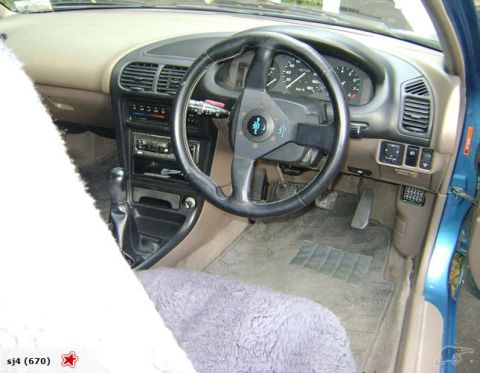 1993 Mazda MX-3 - The interior of a 1993 Mazda MX-3 shows an aftermarket steering wheel, a manual transmission, and a fuzzy seat cover.