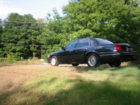 1998 Ford Crown Victoria - A dark 1998 Ford Crown Victoria sedan is parked on a grassy area with trees in the background.