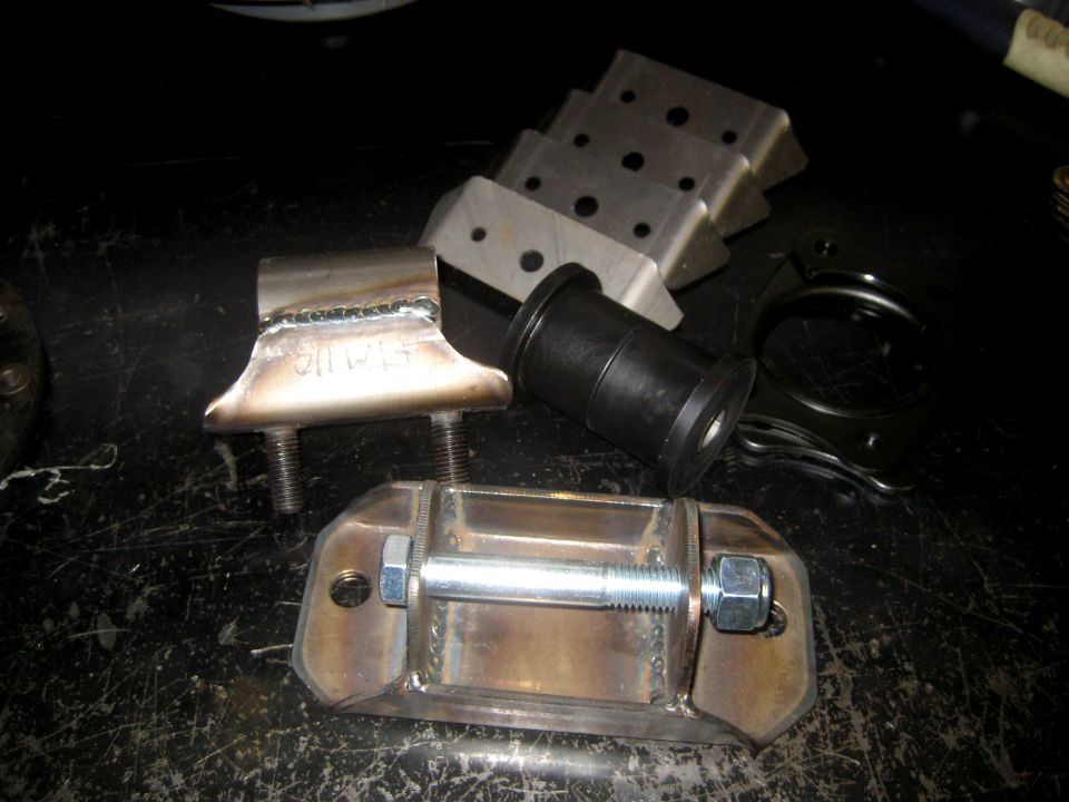Metal brackets and a black rubber bushing on a dark surface. Welded seams are visible on one bracket