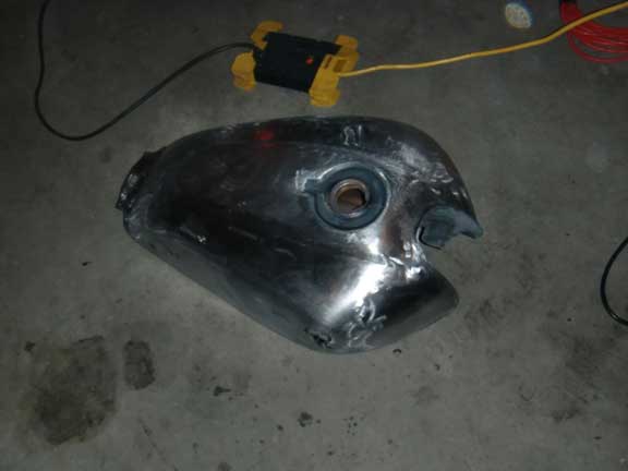 Bare metal motorcycle fuel tank with a central filler cap opening, resting on a concrete floor