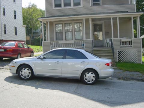 2003 Mazda 6 - A silver sedan is parked on the street in front of a house, with another red car behind it.