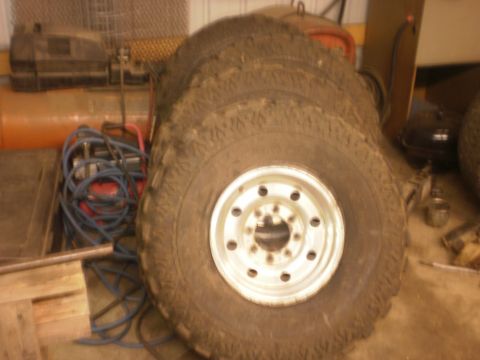 1995 Ford Bronco - Four off-road tires and rims are stacked in a garage next to an air compressor.