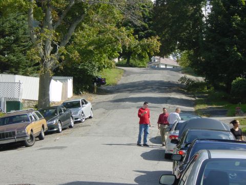 2003 Mazda 6 - Several cars are parked on a street with trees and houses in the background.