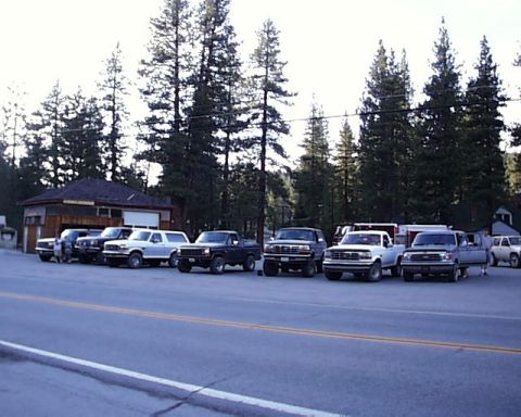 1996 Ford Bronco - A row of trucks and an SUV are parked in front of a building with a wooded area in the background.