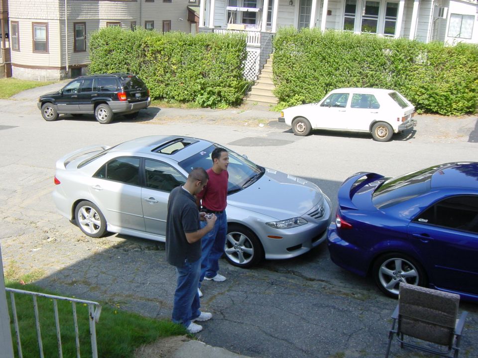 Silver first-gen Mazda 6 sedan at a driveway car meet with two men beside it and a blue Mazda 6 nearby