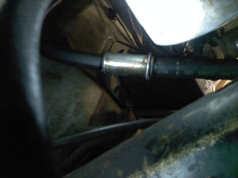1993 Ford Taurus - Dim, tight shot under a vehicle showing a black rubber hose joined to a printed hose by a metal splice or barbed union, running past a dirt…