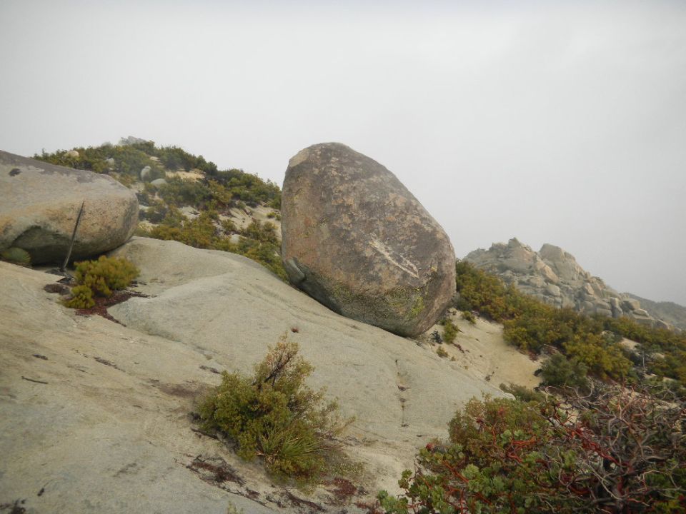 Large boulder resting on a rocky slope with sparse vegetation under a hazy sky