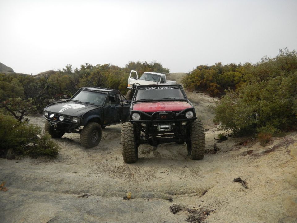 Three modified off-road trucks parked on a dirt trail with scrub brush and rocks