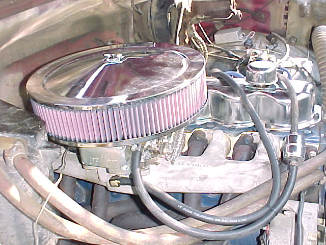 Chrome oval air cleaner on a carburetor with intake manifold and plug wires visible