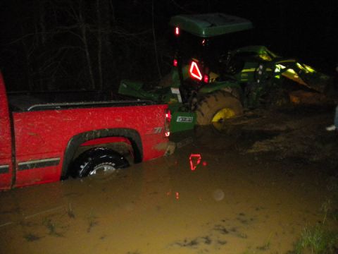 1989 Ford Bronco - A red pickup truck and a green tractor are stuck in deep mud at night.