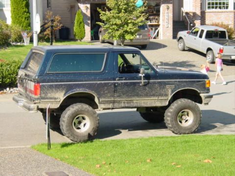 1990 Ford Bronco - A dark blue two-door Ford Bronco SUV with a lift kit and large off-road mud tires, splattered with dried mud, parked in profile on a drivew…