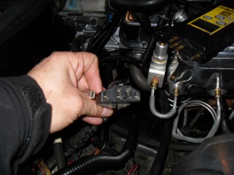 1990 Ford Bronco - A hand holds a multi-pin electrical connector in an engine bay, with metal fuel or brake lines, a wiring harness, and a yellow-labeled comp…