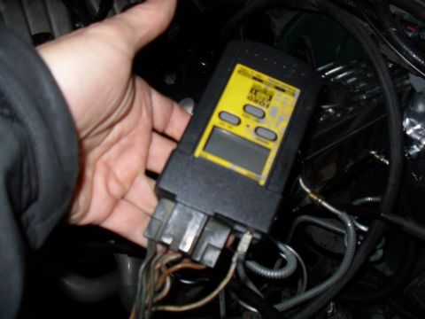 1990 Ford Bronco - A hand holds a black-and-yellow handheld Ford code reader with an LCD screen and three gray buttons, plugged into the vehicle's diagnostic…