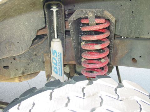 1990 Ford Bronco - Close-up underside view of a lifted suspension showing a red coil spring and a white BDS Suspension shock absorber mounted to a rusty frame…