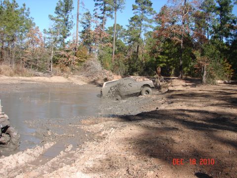 1987 Ford Bronco - An off-road vehicle is stuck in a muddy pit, with trees and a dirt track visible in the background.