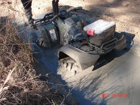 1987 Ford Bronco - An ATV is deeply stuck in thick mud, with a person standing nearby in tall boots.