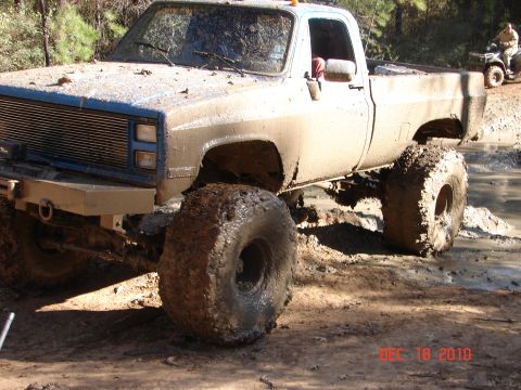 1987 Ford Bronco - A heavily mud-splattered pickup truck navigates a challenging off-road terrain, with an ATV in the distance.