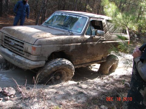 1987 Ford Bronco - A mud-covered 1987 Ford Bronco is stuck in a muddy off-road trail, with a person standing in the background.