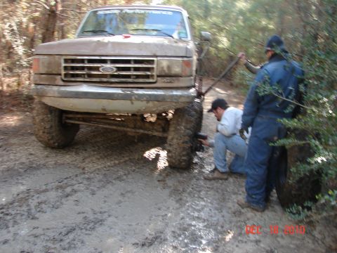 1987 Ford Bronco - A muddy Ford truck is stuck in the woods while two people attempt to fix a tire.