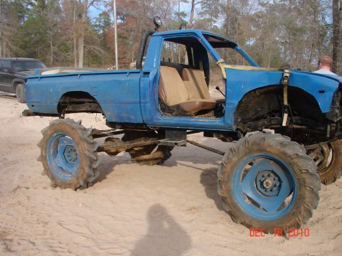1987 Ford Bronco - A heavily modified blue pickup truck with large, aggressive tires is parked on a dirt lot.